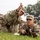 2026 Paralegal Warrior A military training exercise is underway on a grassy field, where an instructor in U.S. Army OCP (Operational Camouflage Pattern) uniform is coaching a soldier in the prone position holding a rifle. The instructor appears to be gesturing with their hand to direct the trainee's aim or attention toward a target. Additional soldiers in uniform are visible in the background observing or awaiting their turn.