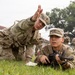 A military training exercise is underway on a grassy field, where an instructor in U.S. Army OCP (Operational Camouflage Pattern) uniform is coaching a soldier in the prone position holding a rifle. The instructor appears to be gesturing with their hand to direct the trainee's aim or attention toward a target. Additional soldiers in uniform are visible in the background observing or awaiting their turn.