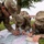 2026 Paralegal Warrior Three U.S. Army soldiers in Operational Camouflage Pattern (OCP) uniforms are conducting map reading and land navigation training outdoors. They are gathered around a large topographic map spread on the grass, with one soldier taking notes in a spiral notebook. The setting appears to be a military installation based on the visible buildings and organized training environment.