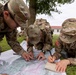 Three U.S. Army soldiers in Operational Camouflage Pattern (OCP) uniforms are conducting map reading and land navigation training outdoors. They are gathered around a large topographic map spread on the grass, with one soldier taking notes in a spiral notebook. The setting appears to be a military installation based on the visible buildings and organized training environment.