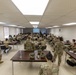 A large group of military personnel in camouflage uniforms (OCP/MultiCam pattern) are attending a briefing or training session in a classroom-style setting. The soldiers are seated at rows of tables facing a projection screen at the front of the room where a presenter is standing. The setting appears to be a military installation classroom or conference room.