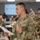 2026 Paralegal Warrior A U.S. Army soldier in Operational Camouflage Pattern (OCP) uniform is presenting or reading from a document in a military classroom or briefing room. The soldier's uniform displays 'U.S. ARMY' tape, a name tape, and a shoulder patch featuring a cactus design, indicating assignment to the 1st Cavalry Division or a Southwest-based unit. Multiple other soldiers in uniform are seated at tables in the background, suggesting a training or briefing event.
