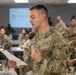 A U.S. Army soldier in Operational Camouflage Pattern (OCP) uniform is presenting or reading from a document in a military classroom or briefing room. The soldier's uniform displays 'U.S. ARMY' tape, a name tape, and a shoulder patch featuring a cactus design, indicating assignment to the 1st Cavalry Division or a Southwest-based unit. Multiple other soldiers in uniform are seated at tables in the background, suggesting a training or briefing event.