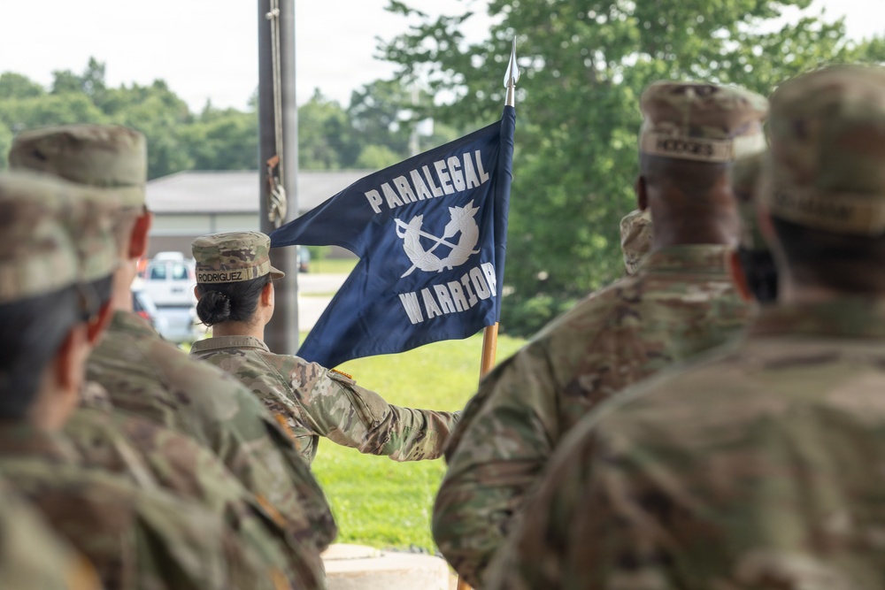 A group of military personnel in U.S. Army OCP (Operational Camouflage Pattern) uniforms are gathered at what appears to be an outdoor ceremony. A soldier identified by nametape as 'RODRIGUEZ' is holding a dark navy blue guidon flag reading 'PARALEGAL WARRIORS' with a crossed-quill-and-sword emblem. The scene suggests a unit formation, activation, or recognition ceremony.