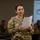 2026 Paralegal Warrior A U.S. Army soldier wearing OCP (Operational Camouflage Pattern) uniform is standing and reading from a sheet of paper, apparently presenting or briefing in a classroom or training environment. Other soldiers are seated in the background, and a U.S. Army presentation slide is visible on a projected screen behind her. The name tape on her uniform partially reads 'OZU'.
