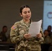 A U.S. Army soldier wearing OCP (Operational Camouflage Pattern) uniform is standing and reading from a sheet of paper, apparently presenting or briefing in a classroom or training environment. Other soldiers are seated in the background, and a U.S. Army presentation slide is visible on a projected screen behind her. The name tape on her uniform partially reads 'OZU'.