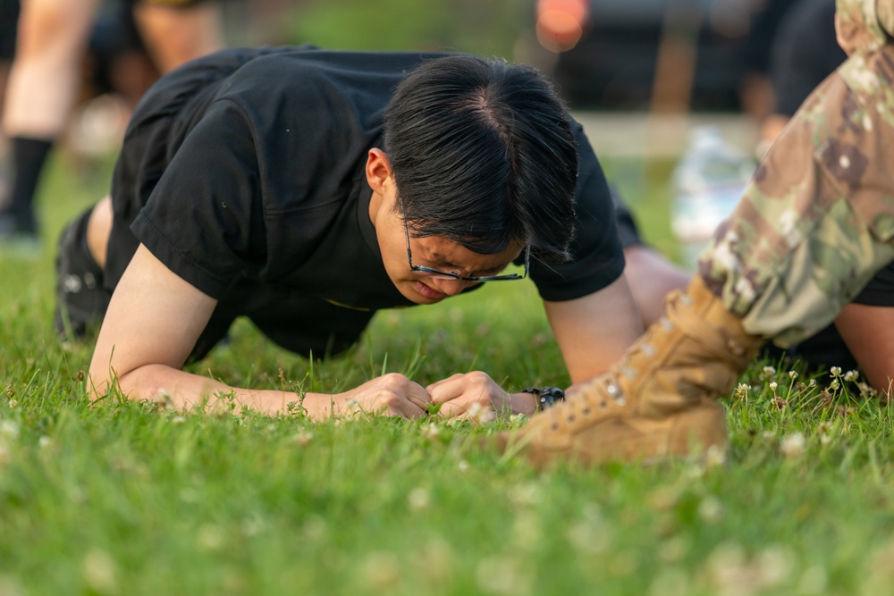 A young person wearing a black t-shirt and glasses is performing a push-up or low plank exercise on a grass field. Their fists are planted on the ground and their body is in a lowered position. In the foreground, another individual wearing military-style camouflage pants and tan combat boots is partially visible.