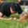 2026 Paralegal Warrior A young person wearing a black t-shirt and glasses is performing a push-up or low plank exercise on a grass field. Their fists are planted on the ground and their body is in a lowered position. In the foreground, another individual wearing military-style camouflage pants and tan combat boots is partially visible.