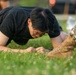 A young person wearing a black t-shirt and glasses is performing a push-up or low plank exercise on a grass field. Their fists are planted on the ground and their body is in a lowered position. In the foreground, another individual wearing military-style camouflage pants and tan combat boots is partially visible.