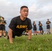 A U.S. Army soldier is performing push-ups on a grass field during what appears to be a physical fitness test or training exercise. Multiple other soldiers in matching black and gold 'ARMY' PT (physical training) uniforms are visible in the background, standing and observing. A clipboard or tablet is partially visible in the upper right corner, suggesting an evaluator is recording performance.