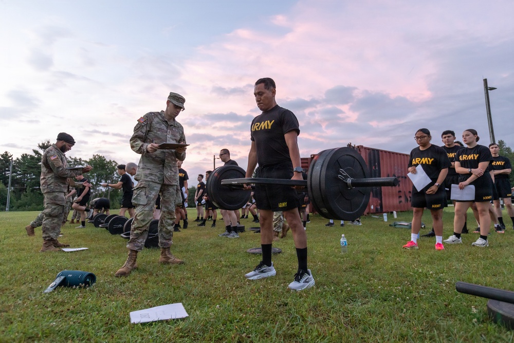 U.S. Army soldiers are conducting a physical fitness assessment, likely the Army Combat Fitness Test (ACFT), on a grass field at dawn or dusk. A soldier in PT (physical training) uniform is performing a deadlift with a loaded barbell while a uniformed evaluator records results on a clipboard. Multiple other soldiers in Army PT gear are observing or waiting their turn in the background.