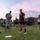 2026 Paralegal Warrior U.S. Army soldiers are conducting a physical fitness assessment, likely the Army Combat Fitness Test (ACFT), on a grass field at dawn or dusk. A soldier in PT (physical training) uniform is performing a deadlift with a loaded barbell while a uniformed evaluator records results on a clipboard. Multiple other soldiers in Army PT gear are observing or waiting their turn in the background.