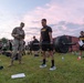 U.S. Army soldiers are conducting a physical fitness assessment, likely the Army Combat Fitness Test (ACFT), on a grass field at dawn or dusk. A soldier in PT (physical training) uniform is performing a deadlift with a loaded barbell while a uniformed evaluator records results on a clipboard. Multiple other soldiers in Army PT gear are observing or waiting their turn in the background.