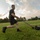 2026 Paralegal Warrior A young muscular man wearing a black Army t-shirt and shorts is performing a sled drag exercise on a grass field during sunset. He is pulling a weighted sled loaded with weight plates using a handle and cable system. The outdoor fitness training is taking place on a well-maintained grassy field with trees and buildings visible in the background.