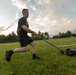 A young muscular man wearing a black Army t-shirt and shorts is performing a sled drag exercise on a grass field during sunset. He is pulling a weighted sled loaded with weight plates using a handle and cable system. The outdoor fitness training is taking place on a well-maintained grassy field with trees and buildings visible in the background.