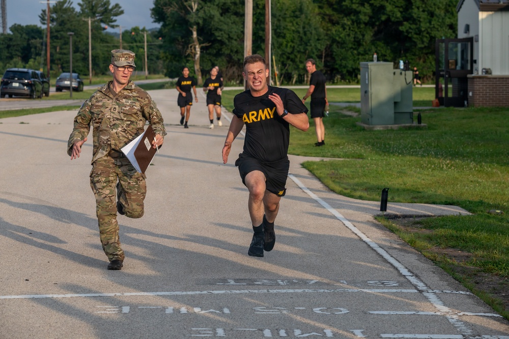 A U.S. Army physical fitness test is underway on a military installation road. A soldier in PT (Physical Training) uniform is sprinting at maximum effort near what appears to be a finish line, while a uniformed evaluator in OCP (Operational Camouflage Pattern) runs alongside holding a clipboard. Additional soldiers in black Army PT uniforms are visible running in the background.