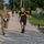 2026 Paralegal Warrior A U.S. Army physical fitness test is underway on a military installation road. A soldier in PT (Physical Training) uniform is sprinting at maximum effort near what appears to be a finish line, while a uniformed evaluator in OCP (Operational Camouflage Pattern) runs alongside holding a clipboard. Additional soldiers in black Army PT uniforms are visible running in the background.