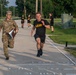 A U.S. Army physical fitness test is underway on a military installation road. A soldier in PT (Physical Training) uniform is sprinting at maximum effort near what appears to be a finish line, while a uniformed evaluator in OCP (Operational Camouflage Pattern) runs alongside holding a clipboard. Additional soldiers in black Army PT uniforms are visible running in the background.