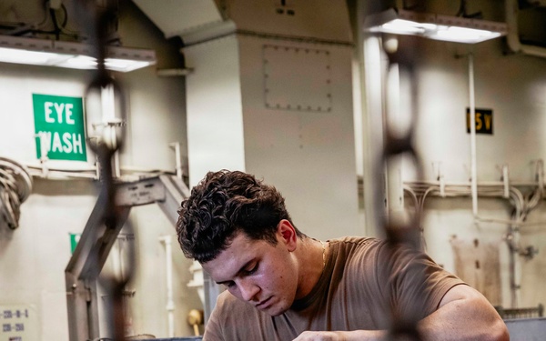 A young male technician in a tan/brown t-shirt is performing maintenance on what appears to be a jet engine or turbine component in an industrial or military workspace. The setting appears to be aboard a naval vessel, indicated by the institutional paint scheme, overhead lighting, and safety signage. The worker is focused intently on manipulating fuel lines or hydraulic connections on the engine casing.