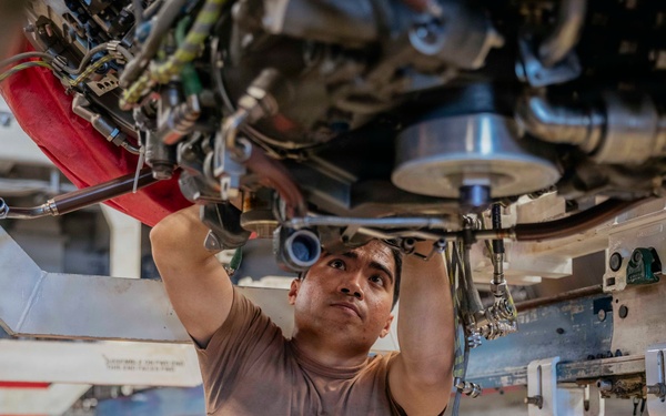 A male technician is performing maintenance work on the underside of what appears to be a military or commercial aircraft, working with both hands raised into the mechanical assembly above him. He is positioned beneath complex machinery including hydraulic lines, structural components, and mechanical systems. The setting appears to be an aircraft hangar, with red equipment and support structures visible in the background.