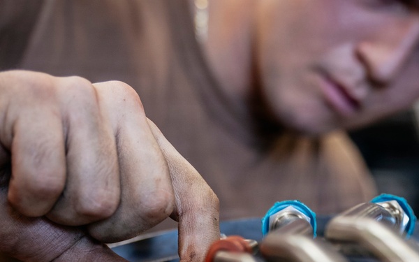 A technician is closely inspecting and working on a complex assembly of metal tubing, fittings, and connectors. The person is using their hands to manipulate or tighten a small fitting or connector within a dense network of pipes and tubes. The equipment appears to be precision mechanical or aerospace hardware based on the high-quality metal components visible.