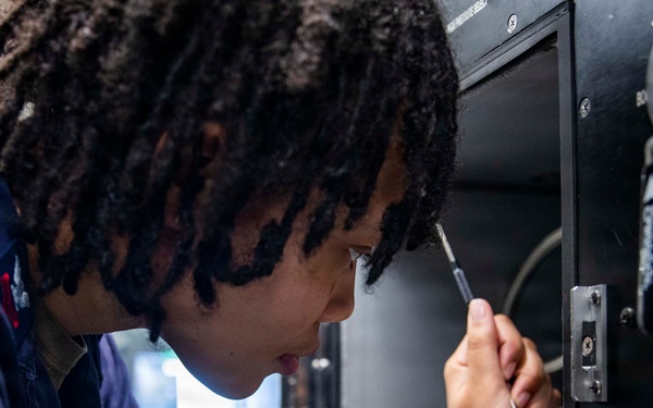A U.S. Navy service member is closely inspecting or working on a piece of electronic or mechanical equipment using a screwdriver. The individual is wearing a Navy coverall uniform with a visible patch and has tattoos on their forearms along with a gold watch. The image captures a focused, hands-on maintenance or repair task in what appears to be a shipboard or technical facility environment.