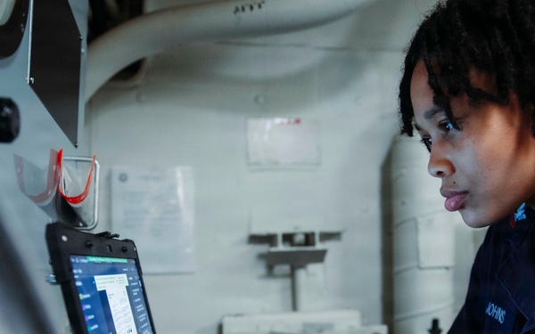 A U.S. military service member, identifiable by a dark navy uniform with 'U.S.' visible on the sleeve and a name tag reading 'JOHNS', is working on a ruggedized laptop computer in what appears to be a confined shipboard compartment. The individual is focused on the laptop screen, which displays what appears to be a software interface or data entry form. The setting suggests an operational or technical workspace aboard a military vessel.