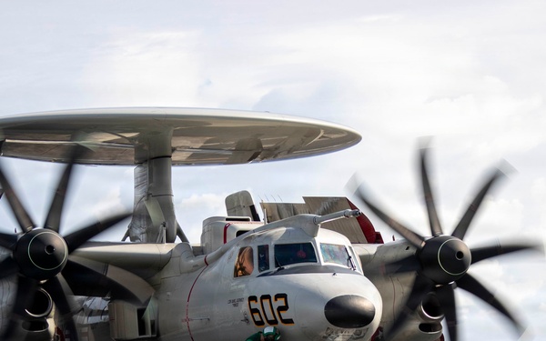 A U.S. Navy E-2 Hawkeye airborne early warning aircraft (bureau number 602) is positioned on the flight deck of an aircraft carrier, with its twin turboprop engines running and rotodome radar disc mounted above the fuselage. Deck crew in yellow and green jerseys are conducting pre-launch operations near the aircraft. The wings appear to be in a folded or transitioning position, and the aircraft is staged near a jet blast deflector.