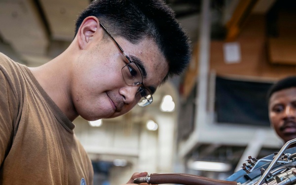 Two individuals in military-style brown t-shirts are working on what appears to be a jet engine or turbine component in an industrial or hangar setting. The foreground technician, wearing glasses, is carefully manipulating fuel lines or hydraulic tubing on the engine assembly. A second technician observes in the background.