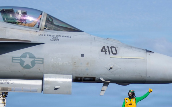 A U.S. Navy F/A-18 Super Hornet (bureau number 410) is positioned on an aircraft carrier flight deck, apparently being prepared for launch. Two flight deck crew members are visible — one in a yellow jersey marked 'FLY 3' and one in a green jersey — performing pre-launch signaling duties. The scene takes place at sea under clear skies.