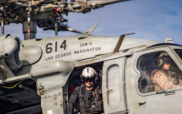 A U.S. Navy helicopter numbered 614, assigned to CVW-5 aboard the USS George Washington, is shown in flight or hovering with crew members visible. The aircraft bears crew name markings including 'LT T.G. ULRICH - BERNIE' and 'AE3 D.J. JOHNSON, BRONX, NY'. Three crew members are visible — one seated in the open cabin door area, one leaning out of the side door, and one in the cockpit.