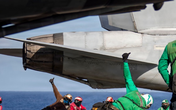 Flight deck crew members are conducting aircraft launch operations on what appears to be a U.S. Navy aircraft carrier. Personnel in green jerseys (catapult and arresting gear officers/shooters) are performing the characteristic launch signal pose, while red-jerseyied crew members crouch nearby. A military jet aircraft, likely an F/A-18 or similar naval aircraft, is positioned on the flight deck ready for catapult launch.