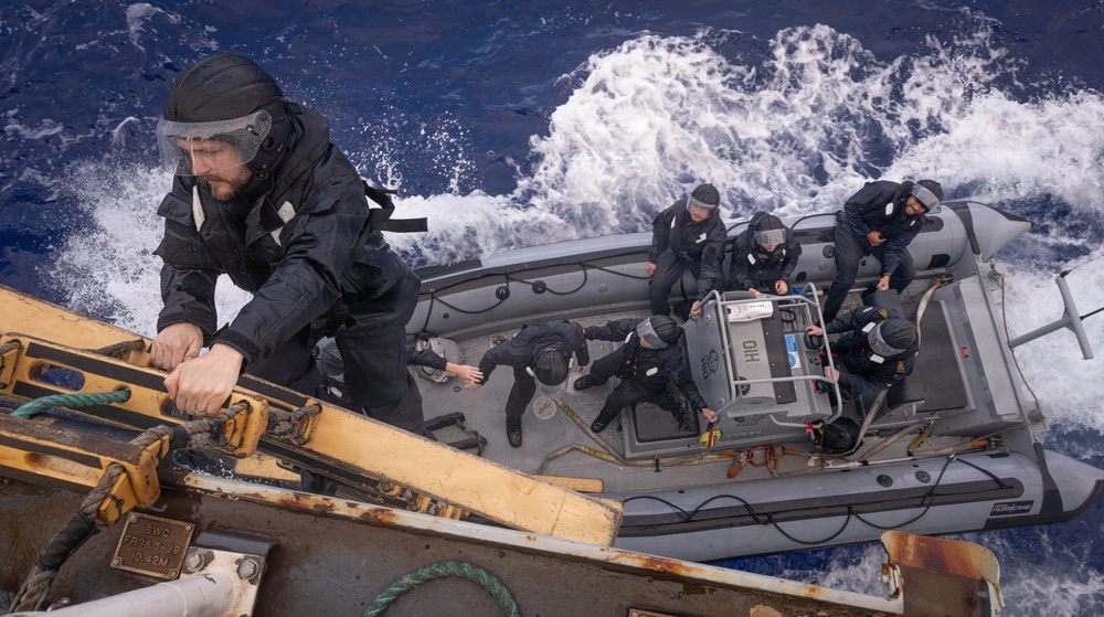 A high-angle photograph captures what appears to be a maritime law enforcement or military boarding operation in rough open ocean conditions. Personnel in dark tactical uniforms and helmets with face shields are aboard a rigid inflatable boat (RIB) alongside a larger vessel. One individual is climbing or transferring between the RIB and the larger vessel's deck.