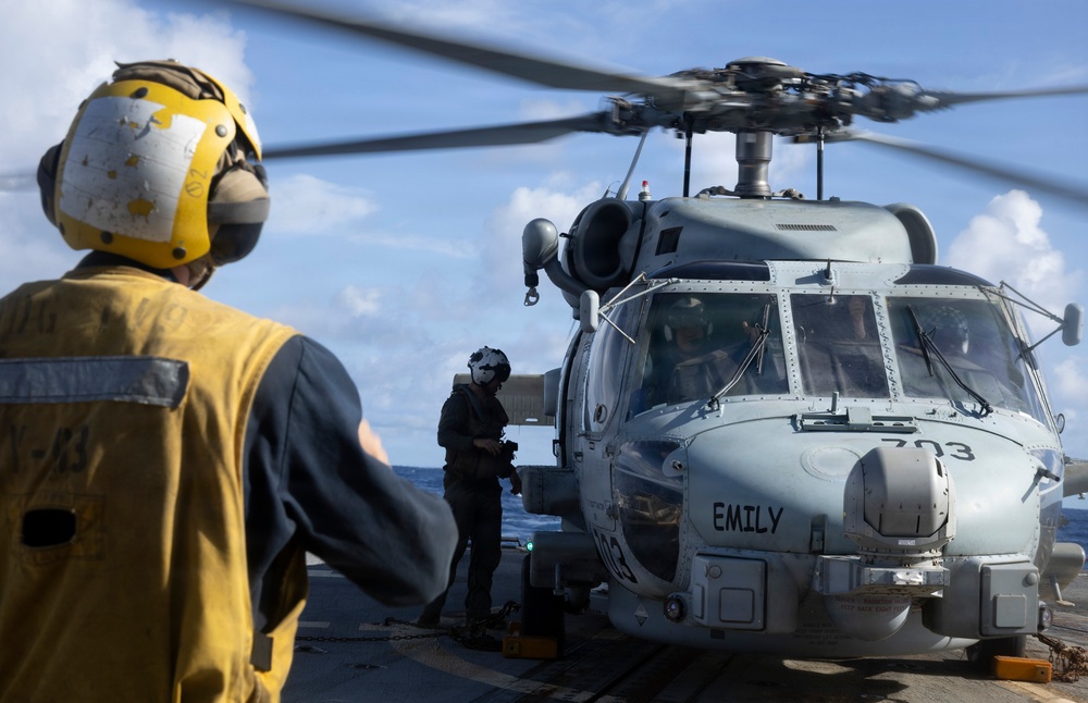 A military helicopter, marked '703' and named 'EMILY', is on the deck of a naval vessel with rotor blades spinning. Deck crew members in protective gear, including a yellow helmet and vest, are visible conducting flight deck operations. The scene takes place at sea under partly cloudy skies.