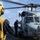 A military helicopter, marked '703' and named 'EMILY', is on the deck of a naval vessel with rotor blades spinning. Deck crew members in protective gear, including a yellow helmet and vest, are visible conducting flight deck operations. The scene takes place at sea under partly cloudy skies.