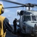 A military helicopter, marked '703' and named 'EMILY', is on the deck of a naval vessel with rotor blades spinning. Deck crew members in protective gear, including a yellow helmet and vest, are visible conducting flight deck operations. The scene takes place at sea under partly cloudy skies.