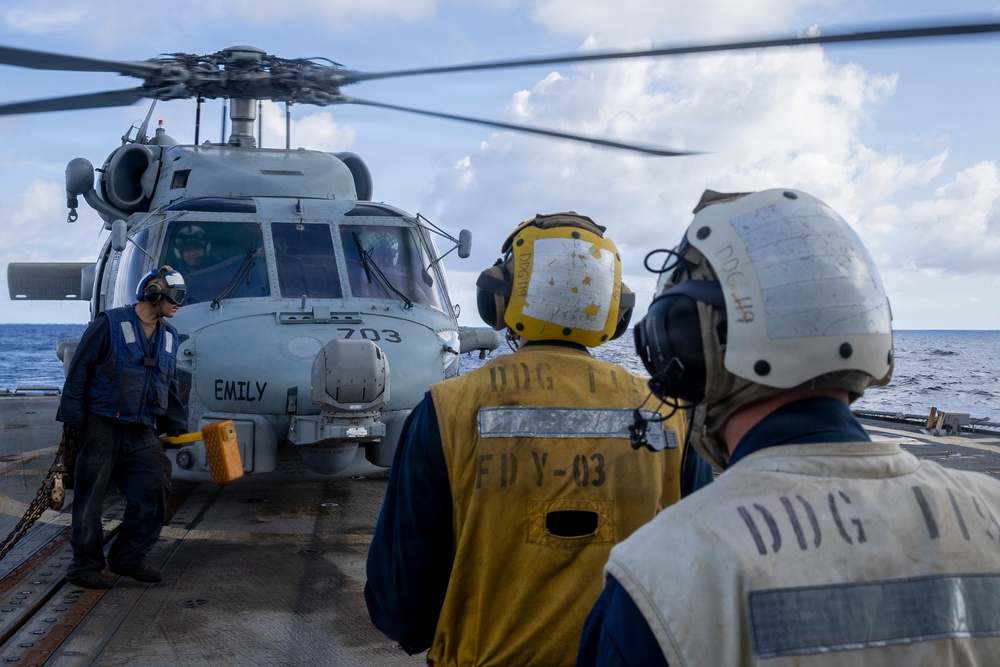 A military helicopter marked '703' and named 'EMILY' is on the flight deck of a U.S. Navy destroyer, identified as DDG-115 based on markings on crew vests and helmets. Three deck crew members are visible, wearing protective helmets and communication headsets, with two in yellow and white vests marked 'DDG 115' and 'FDY-03'. The scene depicts a flight deck operation at sea under partly cloudy skies.