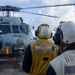 A military helicopter marked '703' and named 'EMILY' is on the flight deck of a U.S. Navy destroyer, identified as DDG-115 based on markings on crew vests and helmets. Three deck crew members are visible, wearing protective helmets and communication headsets, with two in yellow and white vests marked 'DDG 115' and 'FDY-03'. The scene depicts a flight deck operation at sea under partly cloudy skies.