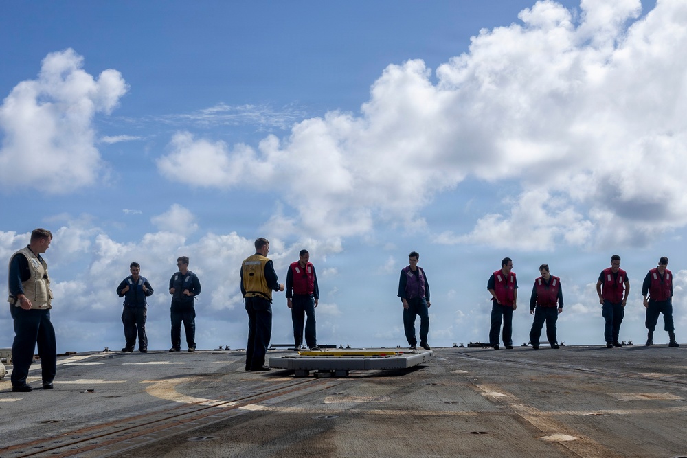 A group of approximately 11 military personnel are standing on what appears to be a naval vessel's flight deck or upper deck. Personnel are wearing various colored safety vests including yellow, red, purple, and tan/white, which are typical color-coded roles on a naval flight deck. The scene appears to be a briefing or muster taking place outdoors under a partly cloudy sky over open water.