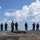 A group of approximately 11 military personnel are standing on what appears to be a naval vessel's flight deck or upper deck. Personnel are wearing various colored safety vests including yellow, red, purple, and tan/white, which are typical color-coded roles on a naval flight deck. The scene appears to be a briefing or muster taking place outdoors under a partly cloudy sky over open water.