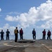 A group of approximately 11 military personnel are standing on what appears to be a naval vessel's flight deck or upper deck. Personnel are wearing various colored safety vests including yellow, red, purple, and tan/white, which are typical color-coded roles on a naval flight deck. The scene appears to be a briefing or muster taking place outdoors under a partly cloudy sky over open water.