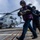 Naval personnel are conducting helicopter refueling operations on the flight deck of a military vessel at sea. Three crew members in colored safety vests and hearing protection are carrying a fuel hose toward a military helicopter marked '703'. The helicopter, a Sikorsky MH-60 Seahawk variant, is parked on the deck with its rotors spinning.