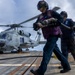 Naval personnel are conducting helicopter refueling operations on the flight deck of a military vessel at sea. Three crew members in colored safety vests and hearing protection are carrying a fuel hose toward a military helicopter marked '703'. The helicopter, a Sikorsky MH-60 Seahawk variant, is parked on the deck with its rotors spinning.