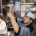 A young male cook or culinary worker wearing a white paper chef's hat and gray uniform is reaching upward to hang or retrieve metal tongs on a utensil rack in a commercial kitchen. The stainless steel environment, organized utensil storage, and food prep materials suggest an active, professional kitchen setting. A tray of raw meat cubes is visible in the foreground on the prep counter.
