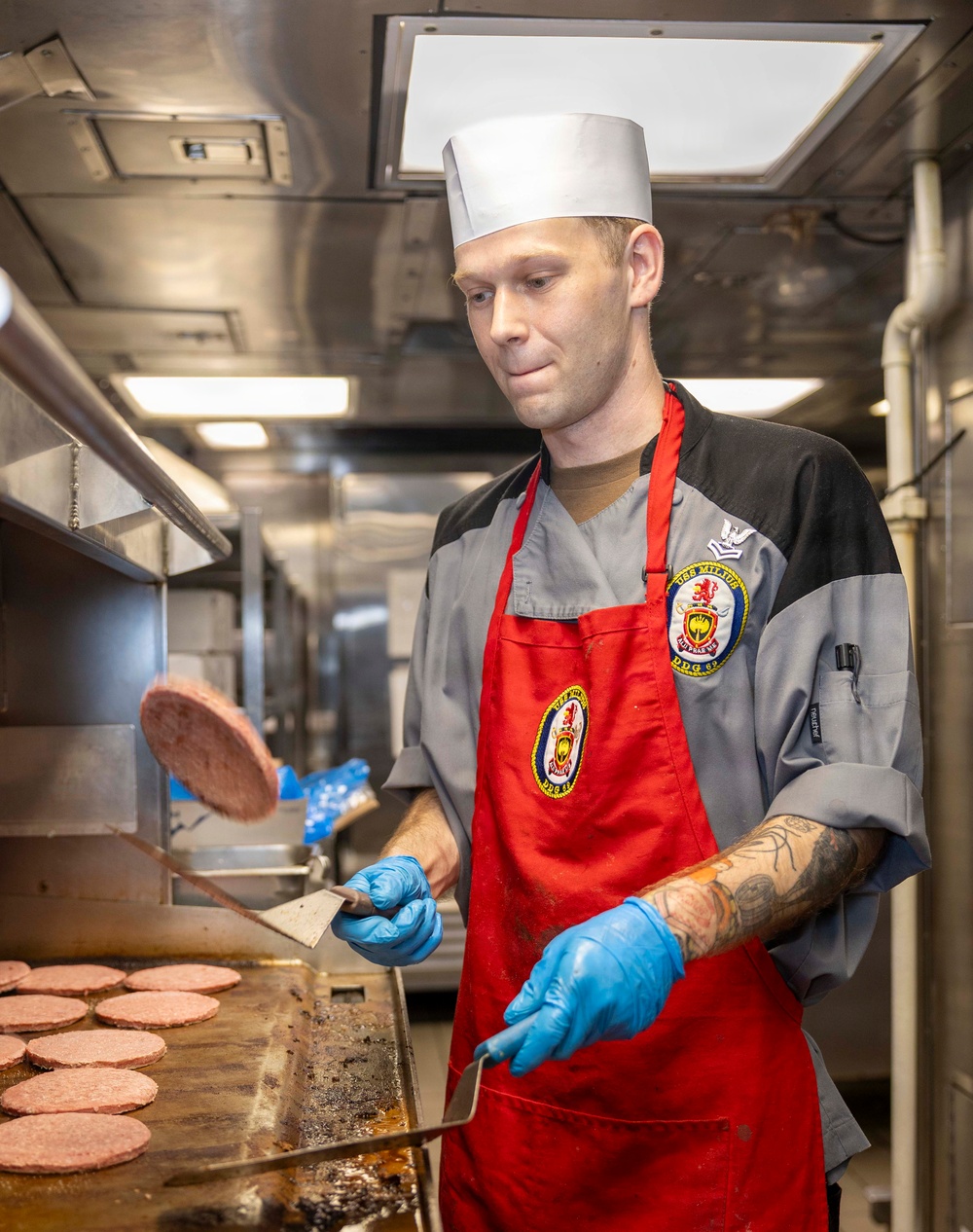 A U.S. Navy culinary specialist is cooking hamburger patties on a large flat-top griddle in a shipboard galley kitchen. He is wearing a white chef's toque, red apron over a gray chef's jacket bearing a ship's patch reading 'USS MILLIUS DDG-69', and blue nitrile gloves. He is in the process of flipping a burger patty mid-air using a metal spatula.