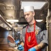 A U.S. Navy culinary specialist is cooking hamburger patties on a large flat-top griddle in a shipboard galley kitchen. He is wearing a white chef's toque, red apron over a gray chef's jacket bearing a ship's patch reading 'USS MILLIUS DDG-69', and blue nitrile gloves. He is in the process of flipping a burger patty mid-air using a metal spatula.