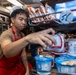 A young male food service worker wearing a red apron and white cap is stocking or organizing food items on a metal rack shelf in what appears to be a military ship's galley or cafeteria. He is handling canned goods and cup-style food containers, including instant noodle cups and cereal cups. A military unit patch is visible on clothing in the background, suggesting a naval vessel setting.