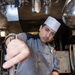 A young male cook or culinary specialist, wearing a white paper chef's hat and gray uniform with a military-style patch, is serving or stirring a large tray of what appears to be baked beans or a similar legume dish in a commercial kitchen. The photo is taken from a low angle close to the food tray, creating a dramatic perspective. The setting appears to be a military galley or institutional kitchen based on the uniform and environment.