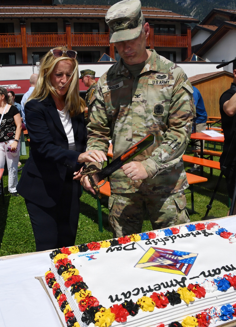 A U.S. Army Colonel named Drouin and a civilian woman are jointly cutting a large ceremonial cake using a military saber. The cake is decorated with German and American colors (black, red, yellow, and blue/red/white) and reads 'German-American Friendship Day'. The event appears to be taking place outdoors at a military community event, likely in Germany based on the Alpine-style architecture visible in the background.