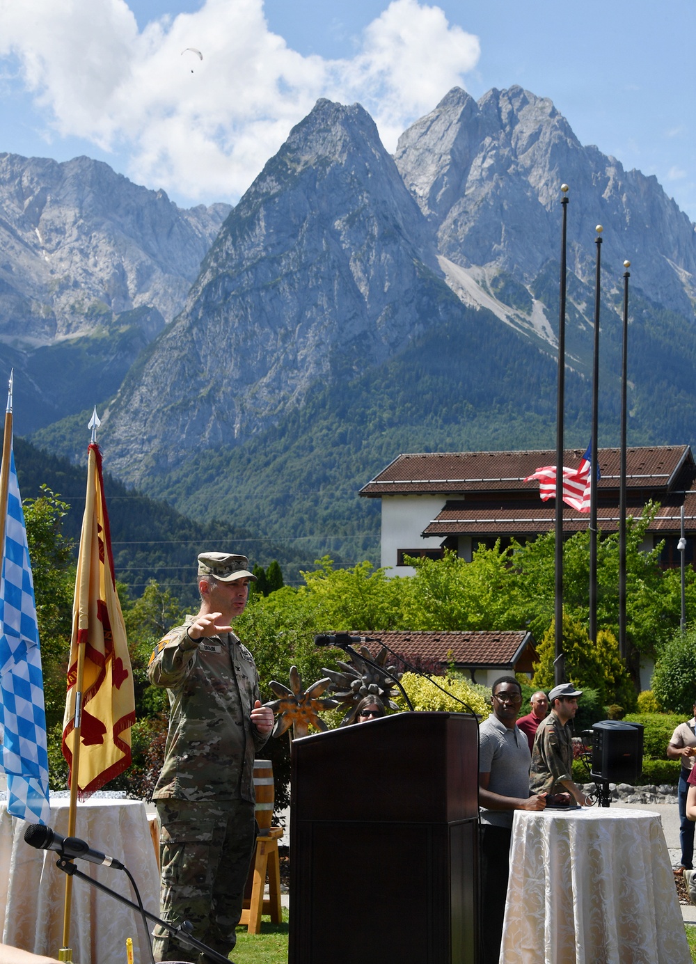 A U.S. Army officer in camouflage uniform speaks at an outdoor podium during what appears to be a military ceremony. The event is set against a dramatic Alpine mountain backdrop with a U.S. flag visible on a flagpole, suggesting a military installation in the Bavarian Alps region of Germany. Multiple ceremonial flags, including a blue-and-white Bavarian flag and a U.S. Army unit flag, are displayed alongside the speaker.